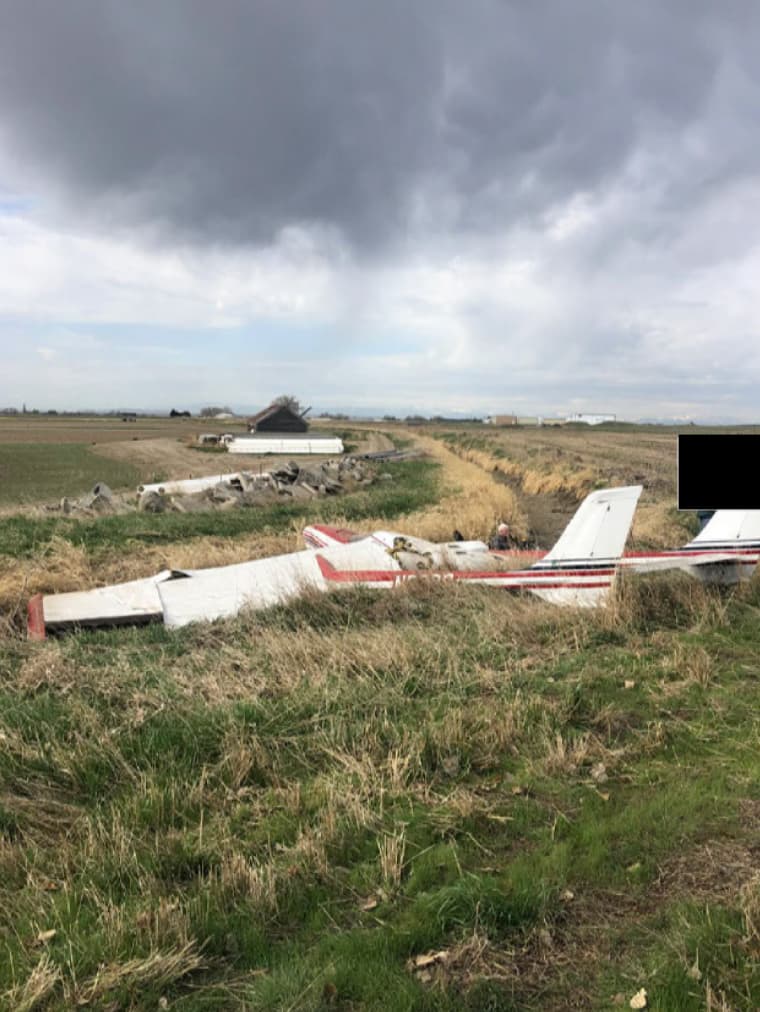Overhead view of a damaged Cessna P337H aircraft N62PC resting in a grassy irrigation ditch under a cloudy sky, with tail section visible and wings crumpled.